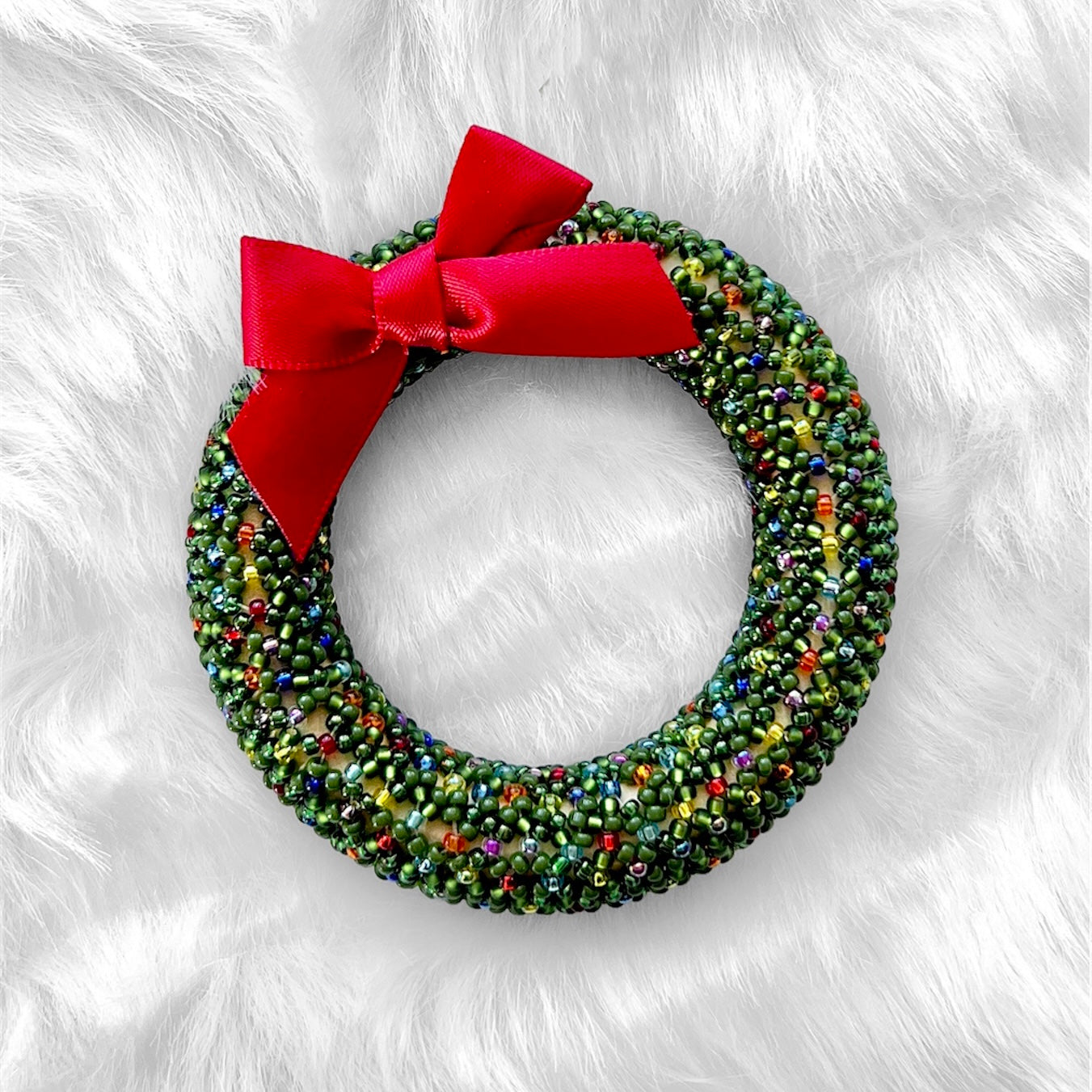 beaded christmas wreath ornament made with green seed beads and a red bow on a white faux fur backdrop