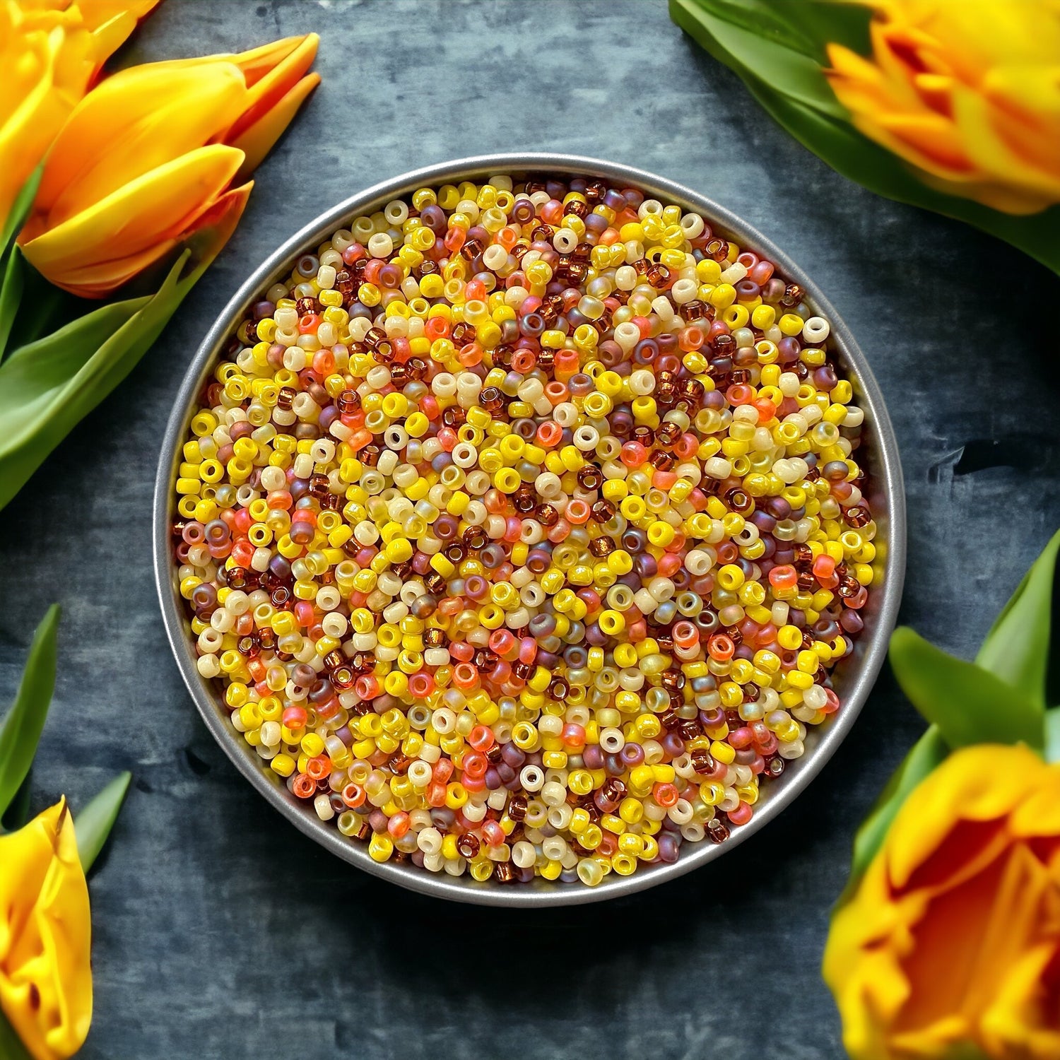 A mix of yellow and orange 11/0 Miyuki seed beads in a silver tray on a dark wood surface surrounded by deep yellow tulips