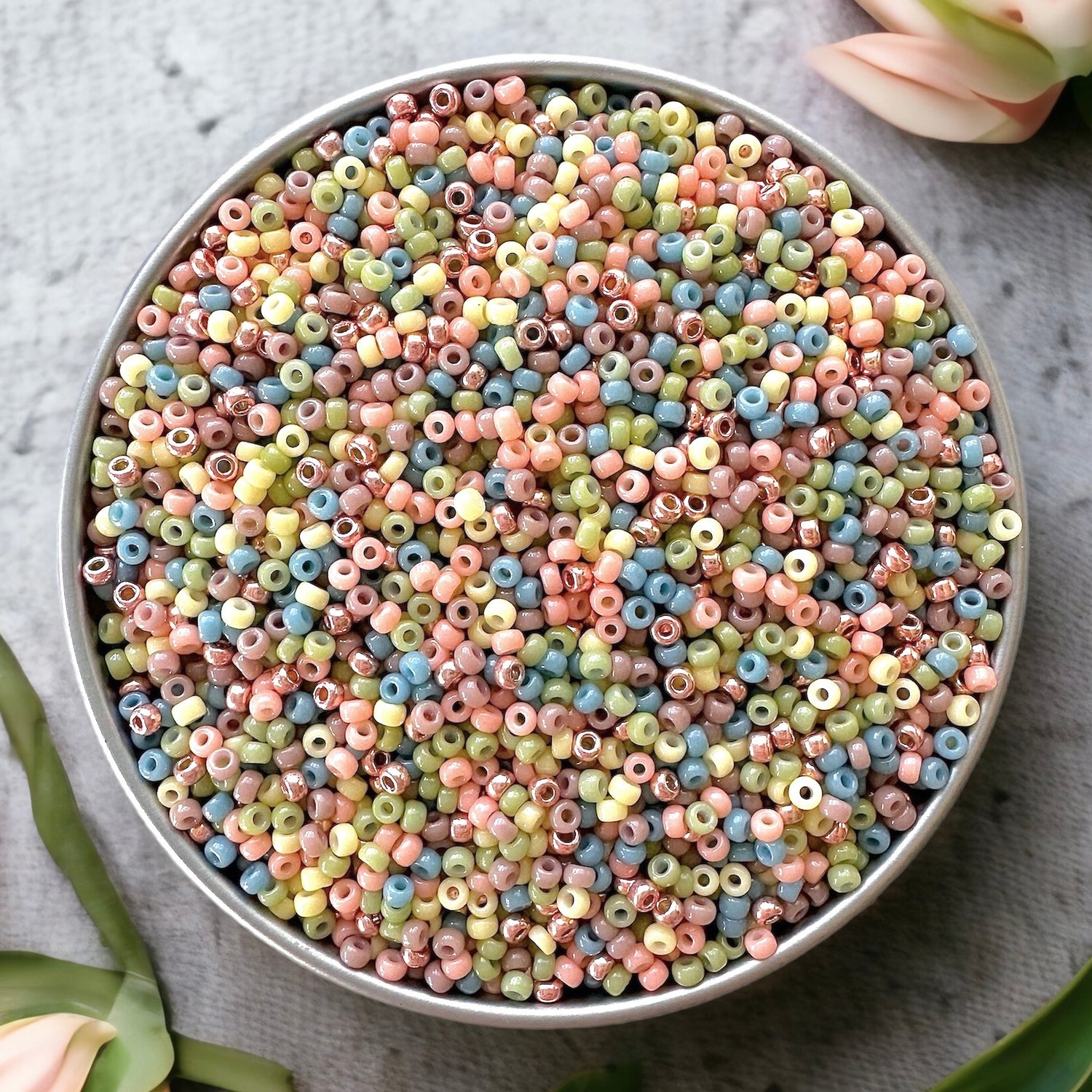 Pastel mix of Miyuki 11/0 seed beads by The Bead featuring pastel shades of peach, yellow, green, blue, and purple in a silver container on a gray concrete surface with peach tulips.