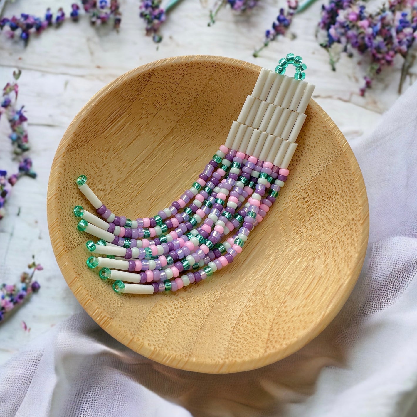 Bugle bead fringe earrings made with cream bugle beads and 11/0 Miyuki seed beads in shades of purple, pink, and teal green in a woodern dish