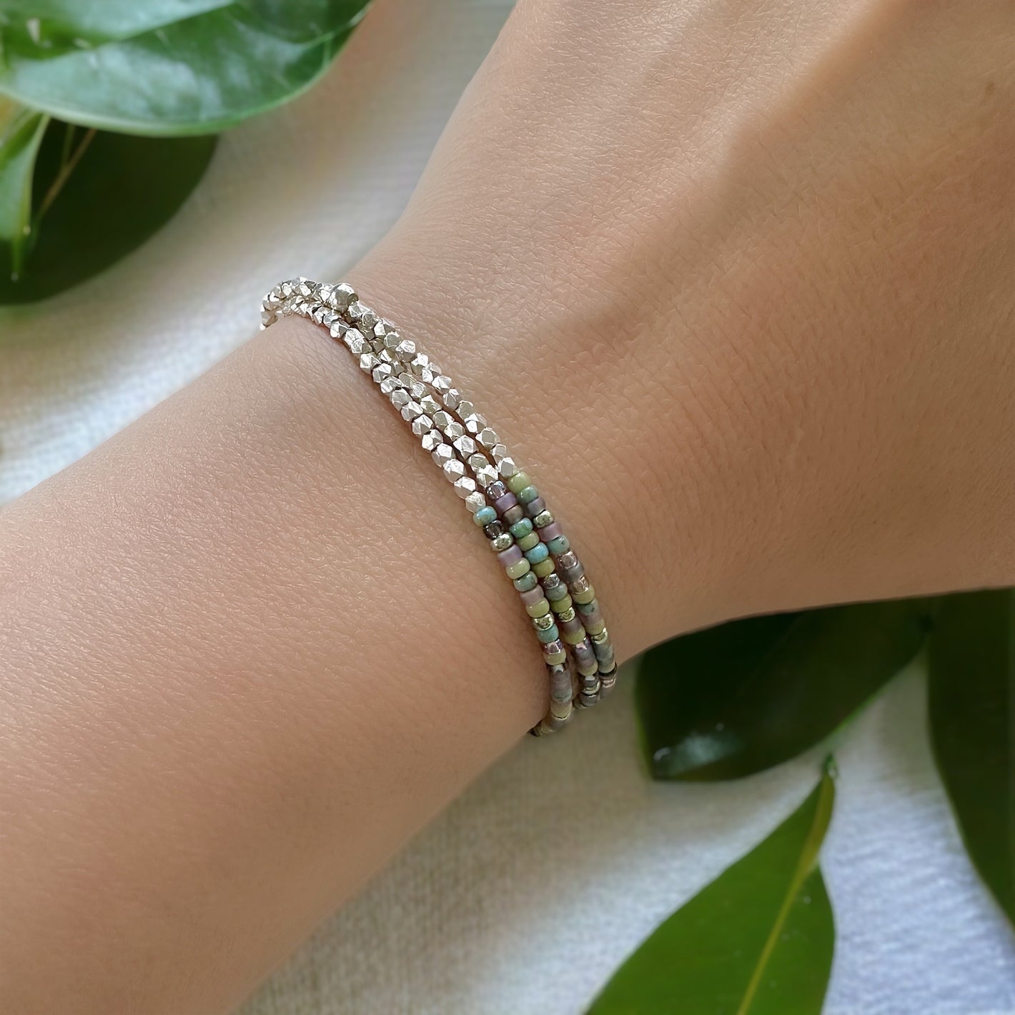 Beaded triple wrap stretch bracelet made with 11/0 Miyuki seed beads and 2mm Karen Hill Tribe fine silver beads worn on a woman's wrist against a white cloth with green leaves 