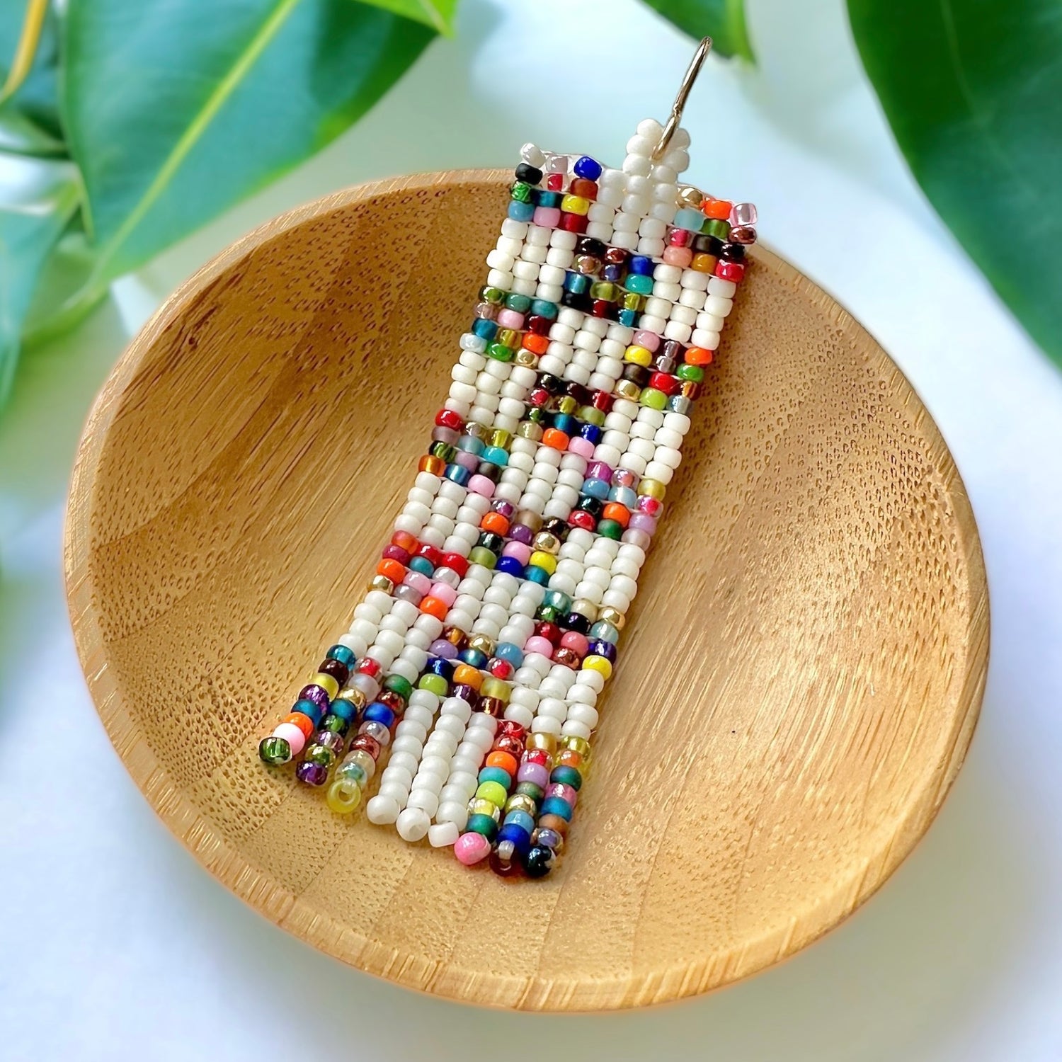 Beaded earrings with a patchwork quilt pattern and mini fringe made with Miyuki 11/0 seed beads in a wooden dish on a white surface surrounded by green leaves