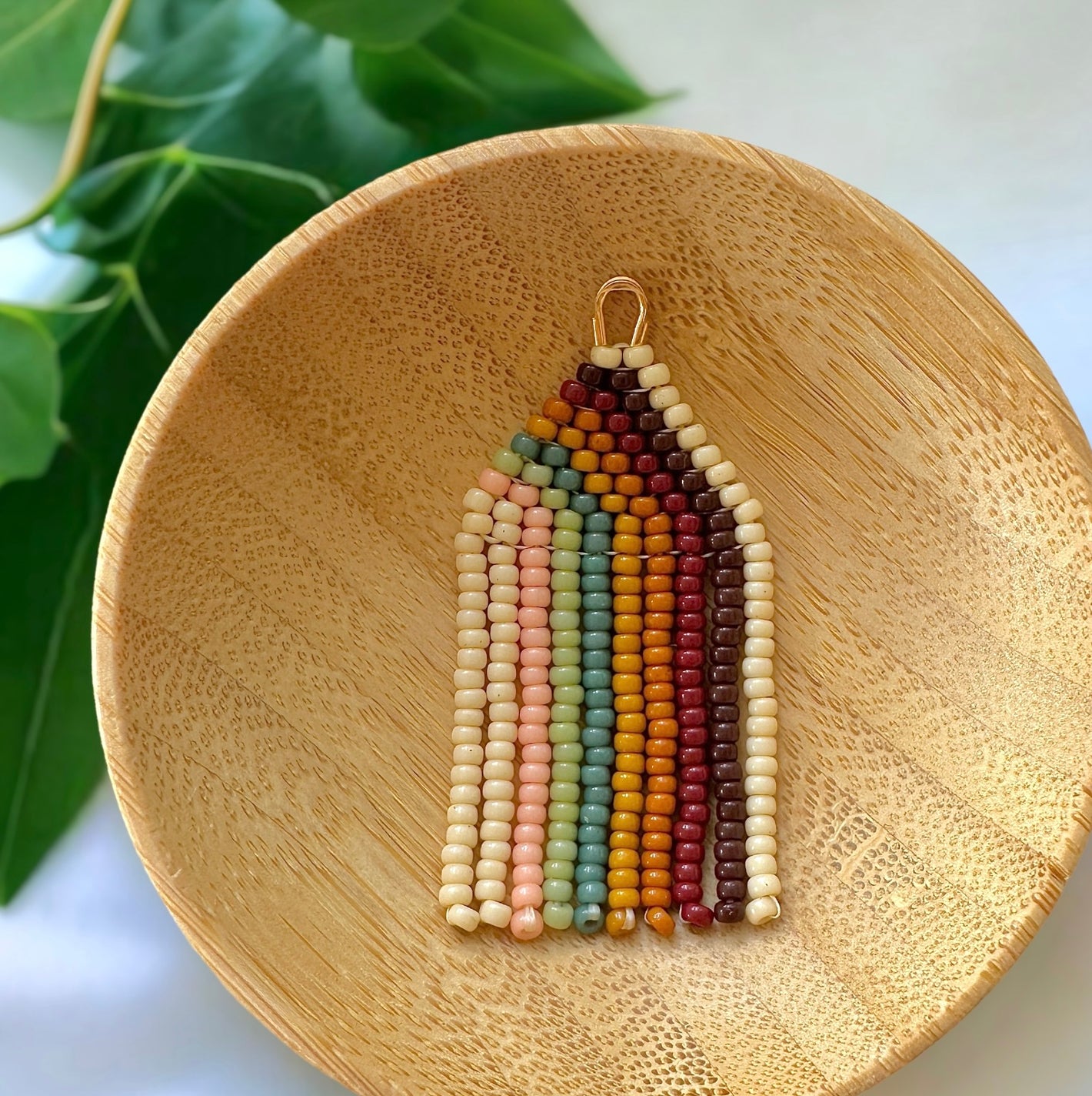 Mini boho rainbow beaded fringe earrings in a wooden bowl on a white cloth surrounded by green leaves
