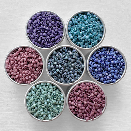 Seven round containers, each filled with Mermaid Miyuki 11/0 Delica Beads in different colors including purple, blue, teal, and pink, arranged neatly on a light gray wooden surface.