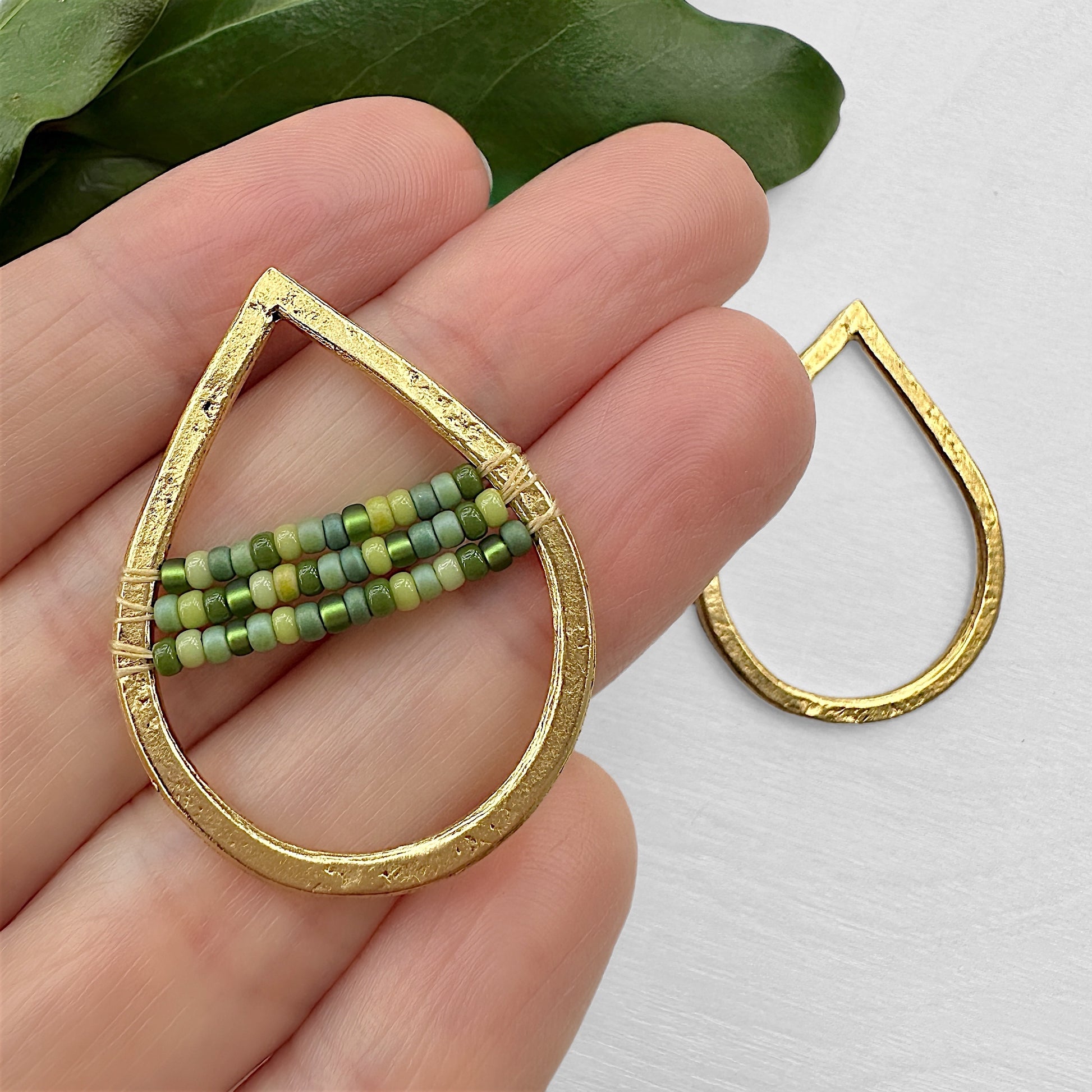Gold teardrop-shaped earrings with Green Magic 11/0 Miyuki Seed Bead Mix accents, displayed on a green leaf against a white background. One earring is filled with beads, and the other is empty.