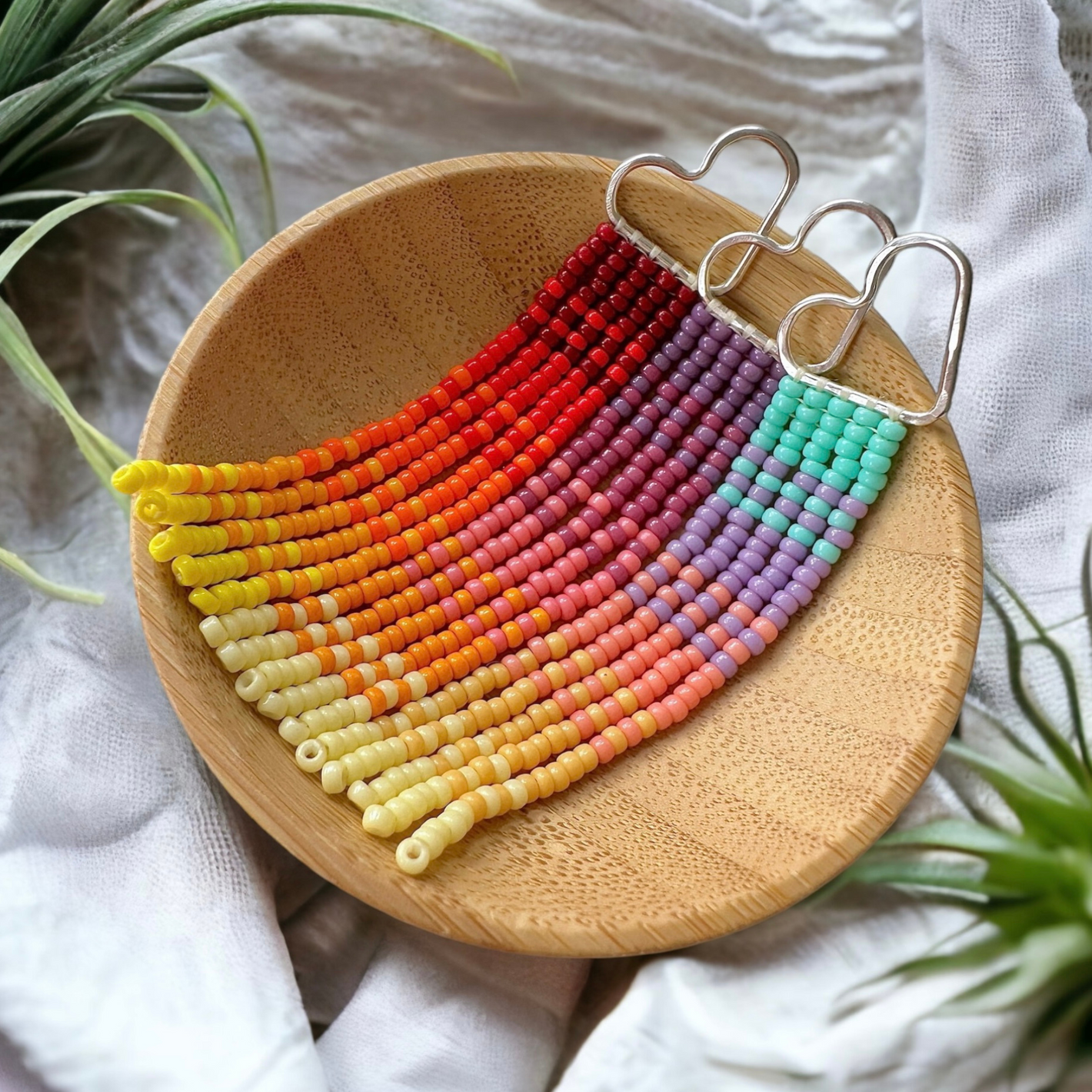 Sunset and sunrise theme beaded fringe earrings on silver heart frames in a wooden dish on a white cloth