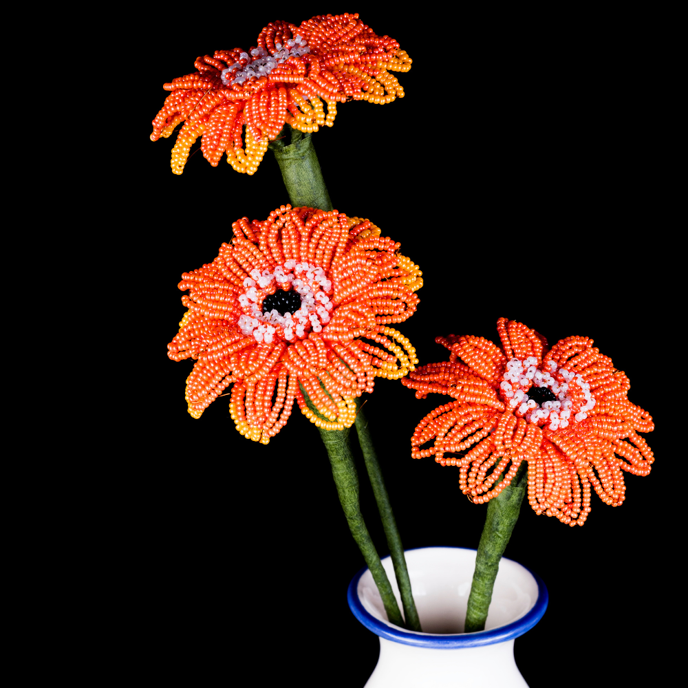 three orange and yellow beaded flowers with green stems in a white vase on a black background