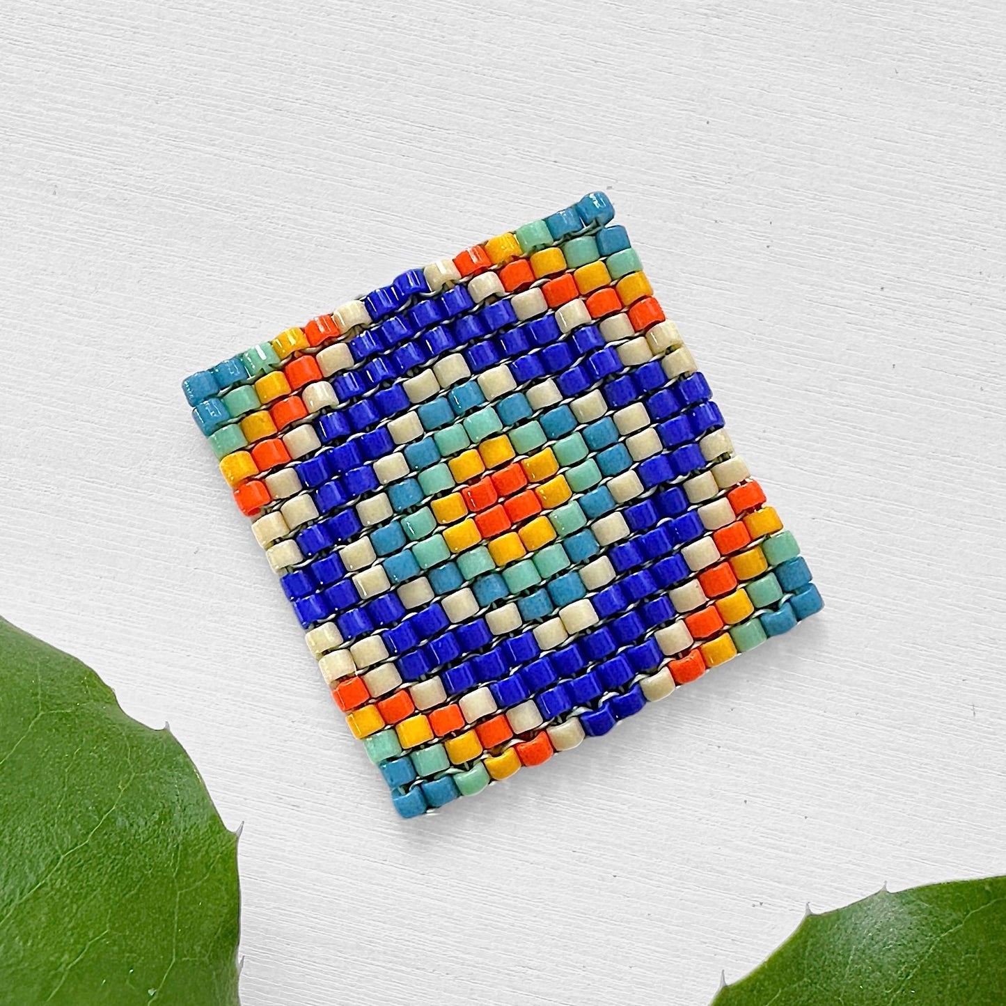 Beaded square made with square stitch and Miyuki seed beads in shades of blue, turquoise, orange, and cream on a white background.