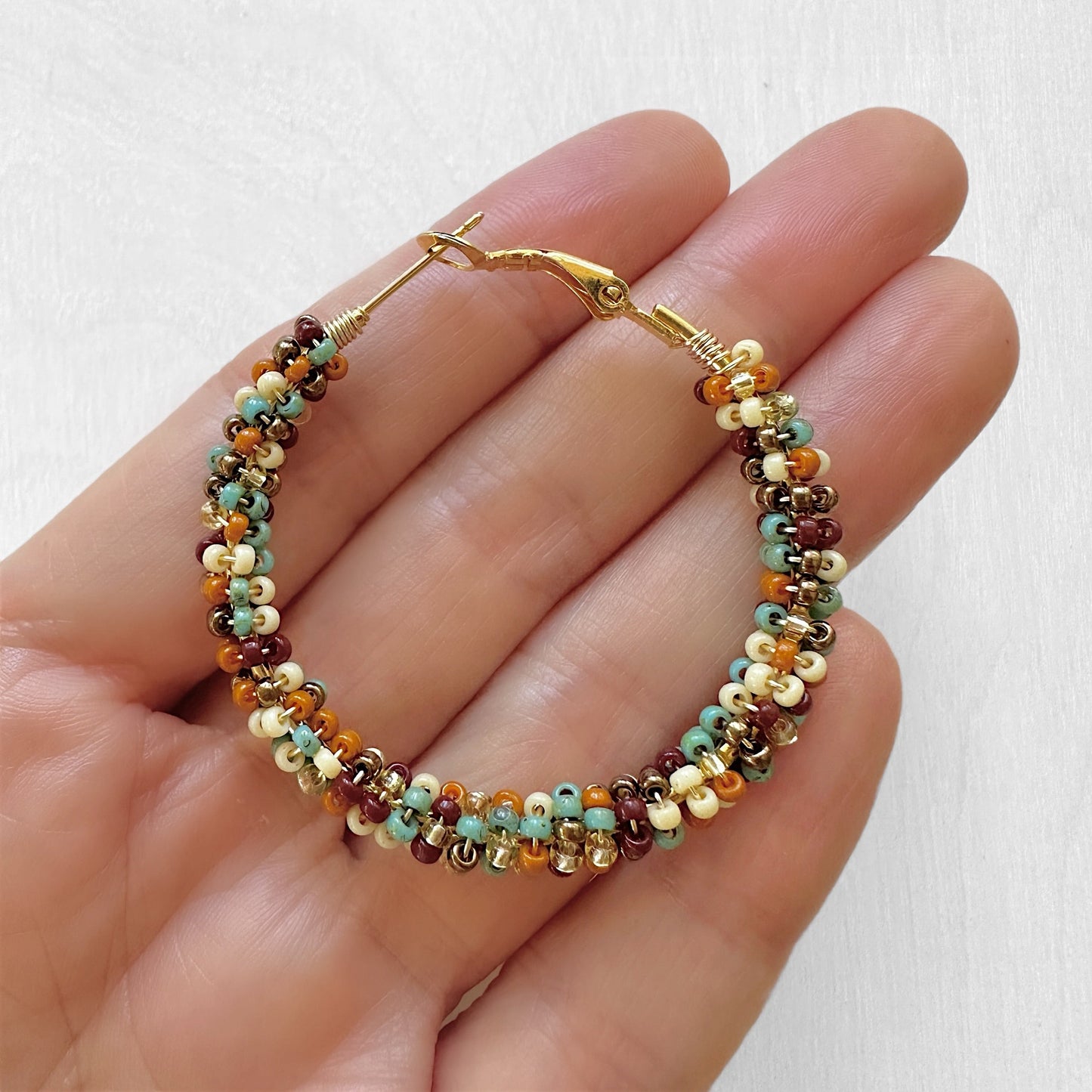 A hand holding a hoop earrings made with Miyuki seed beads in sburgundy, sienna, cream, gold, bronze, and turquoise.