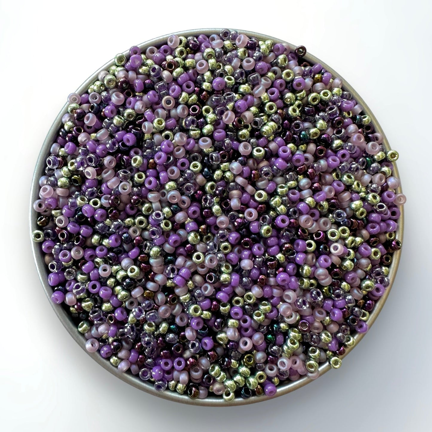 Mix of witch inspired Miyuki 11/0 seed beads, including purple and green in a silver container on a white background