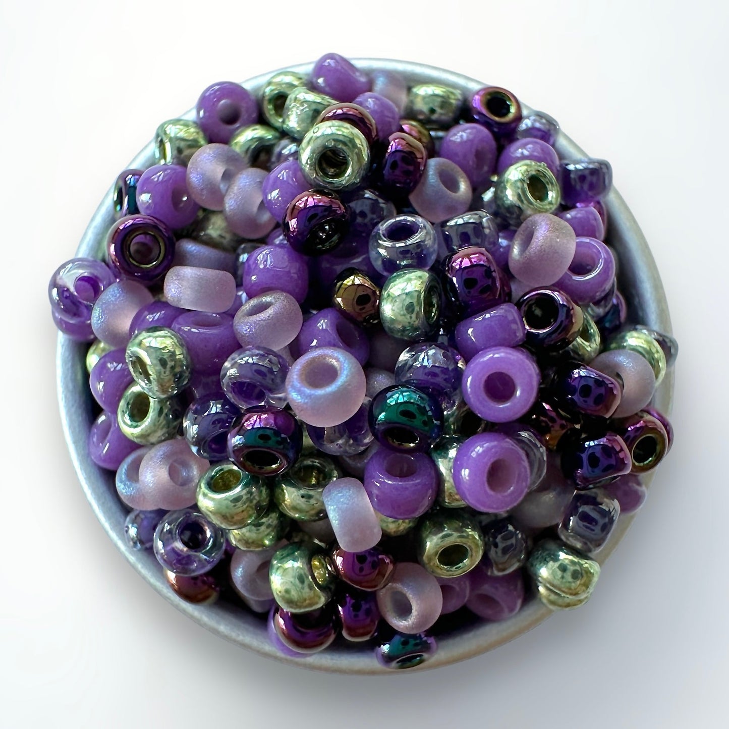 Mix of witch inspired Miyuki 11/0 seed beads, including purple and green in a silver container on a white background