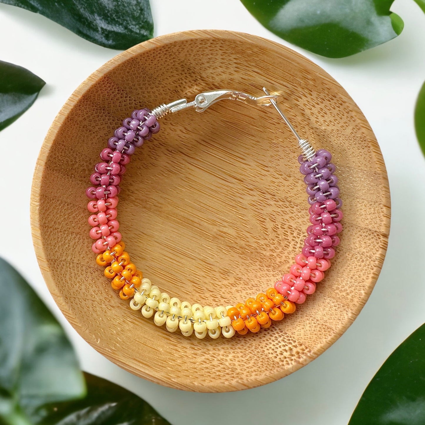 Hoop earring made with Miyuki seed beads in shades of pink, orange, yellow and purple in a wooden bowl.