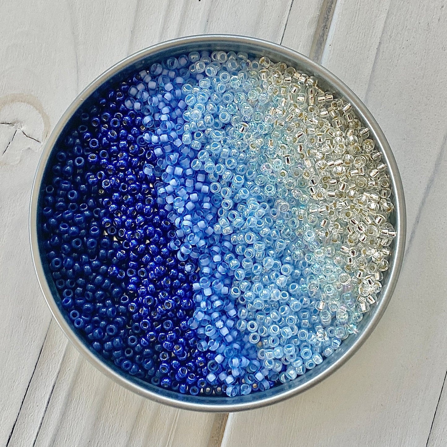 A blue to silver gradient of Miyuki 11/0 seed beads in a silver container on a white wood surface by The Bead Mix