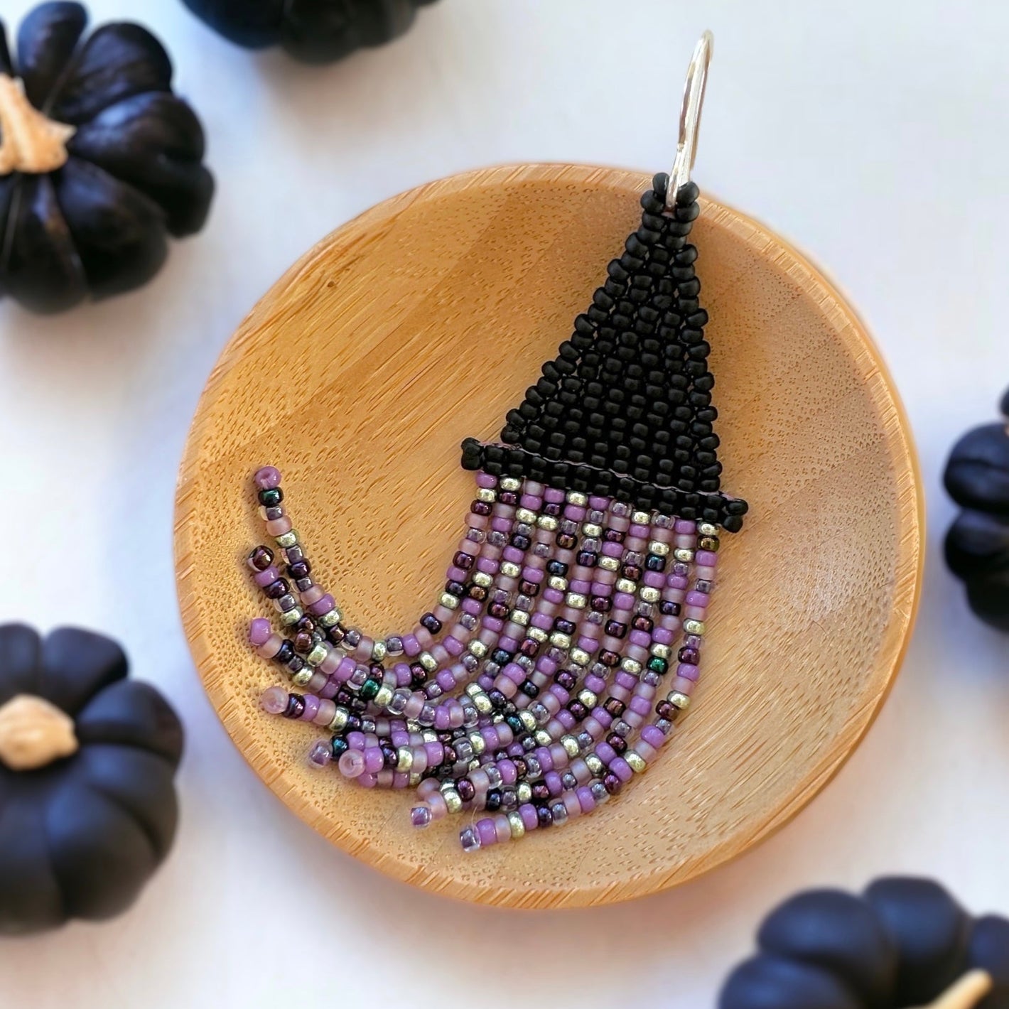 Brick stitch beaded fringe earrings in the shape of a witch's hat with black, purple, and green Miyuki 11/0 seed beads in a wooden dish on a white background surrounded by black pumpkins