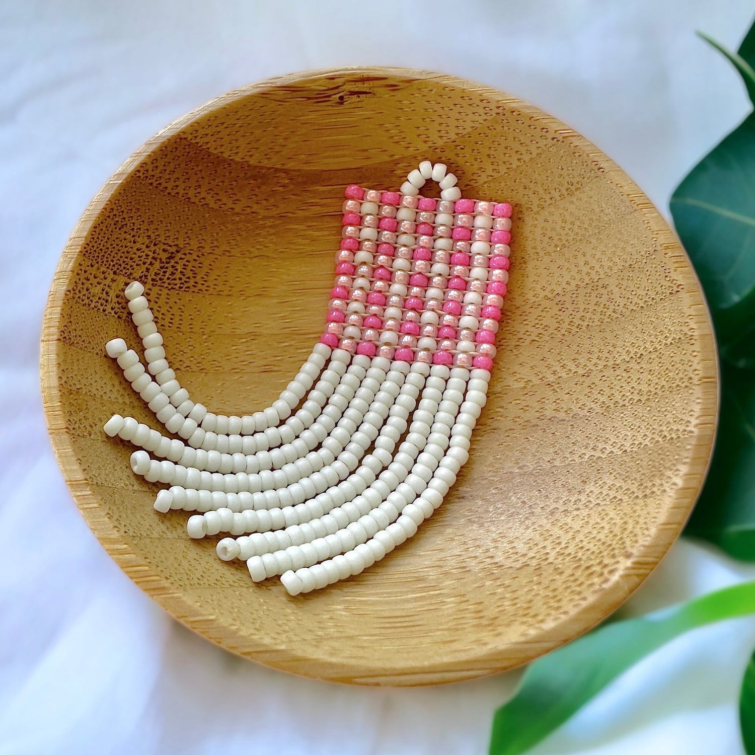 A beaded fringe earring with a pink gingham pattern in a wooden dish on a white cloth