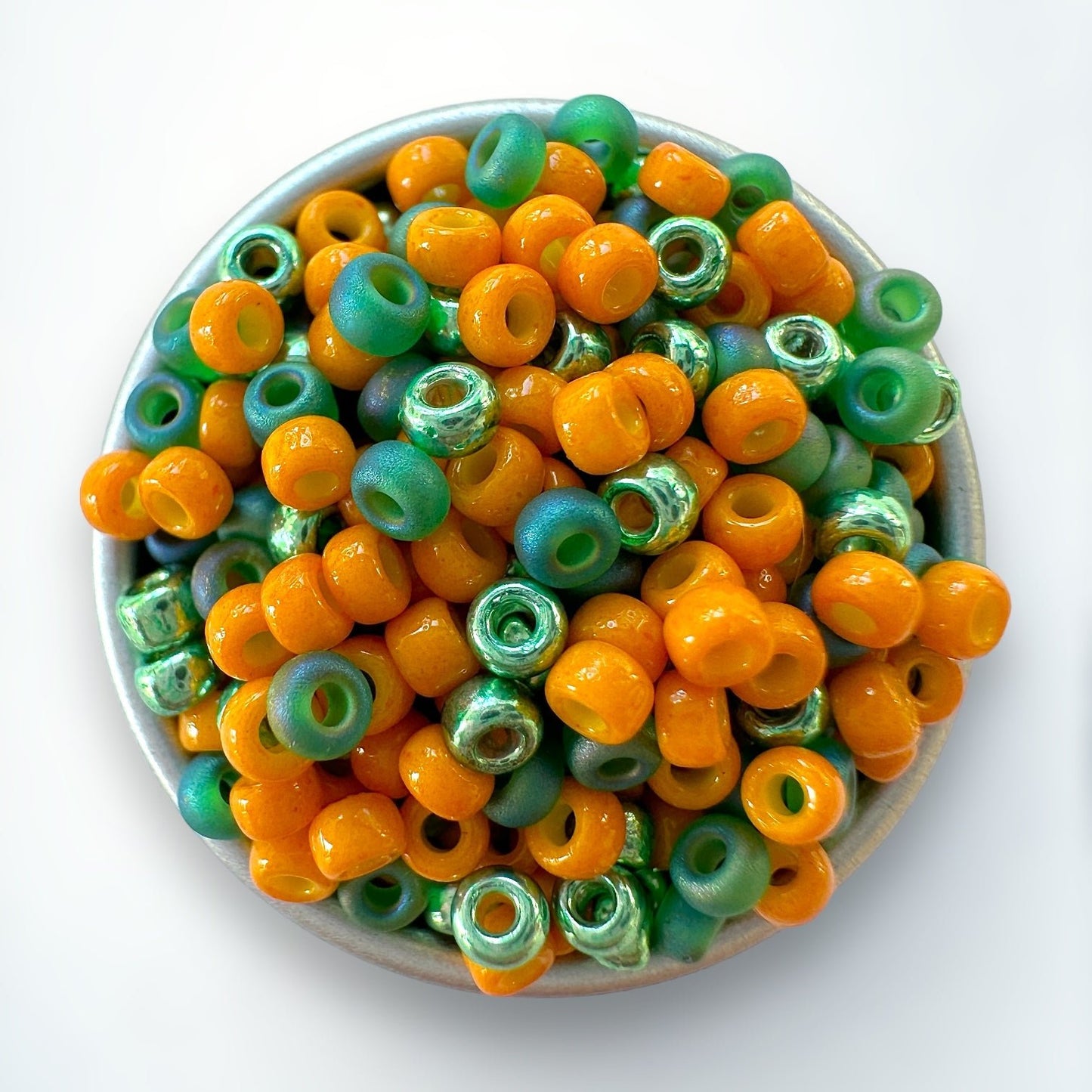 A mix of orange and green Miyuki 11/0 seed beads mixes created by The Bead Mix in a silver container with a white background
