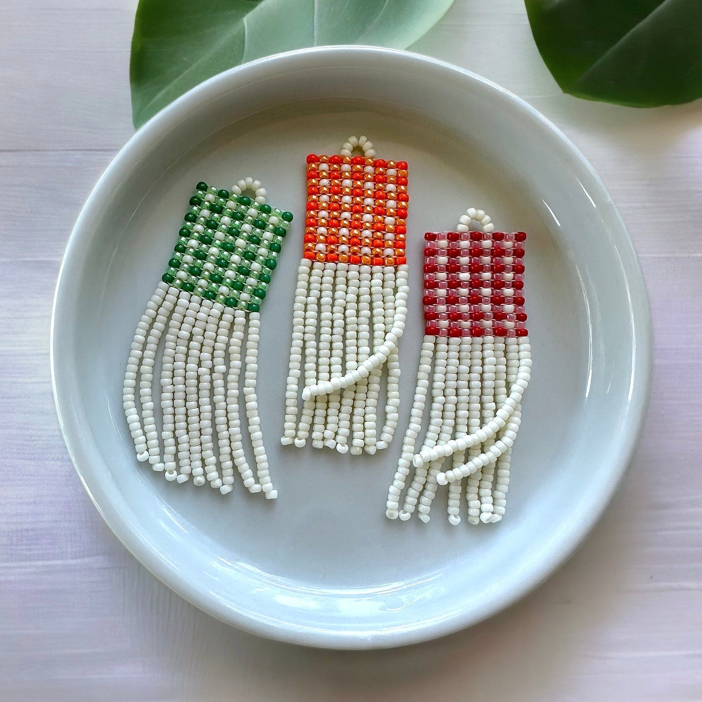 Set of three beaded fringe earrings with a red, green, and orange gingham pattern in white circular ceramic dish on a white cloth background