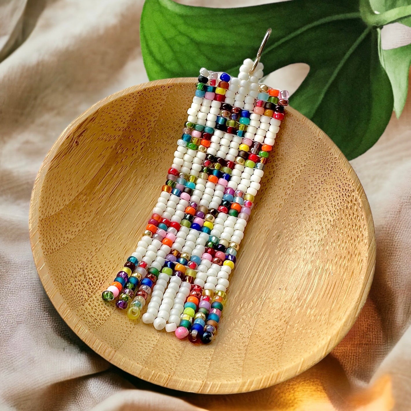 Patchwork quilt beaded earring in a wooden dish surrounded by tan fabric and a green leaf. The earring was made using square flat stitch. It has a gold earring hook and mini fringe.