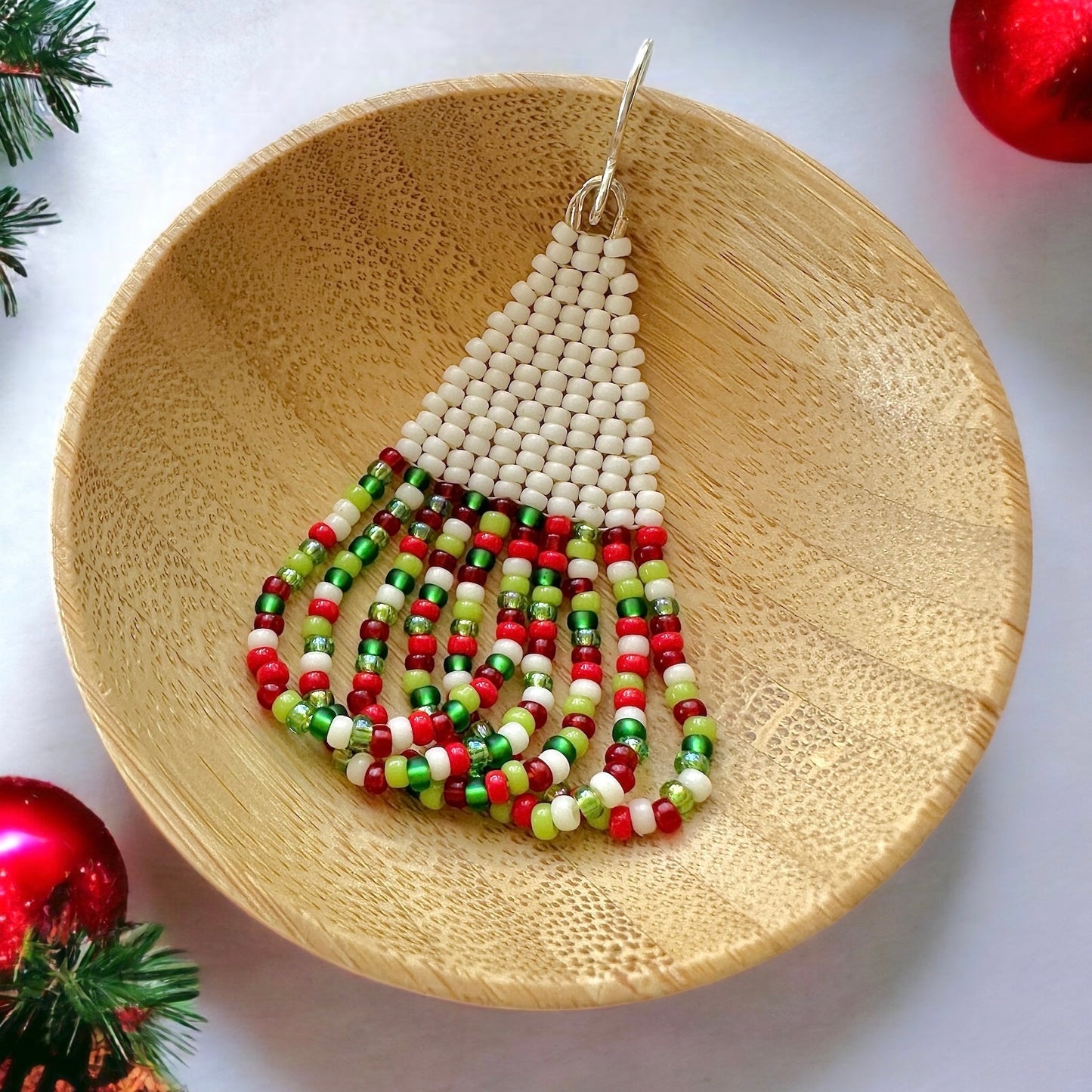 A beaded brick stitch earring with swoop fringe made with cream and christmas colored Miyuki 11/0 seed beads in a wooden dish on a white background surrounded by Christmas decorations