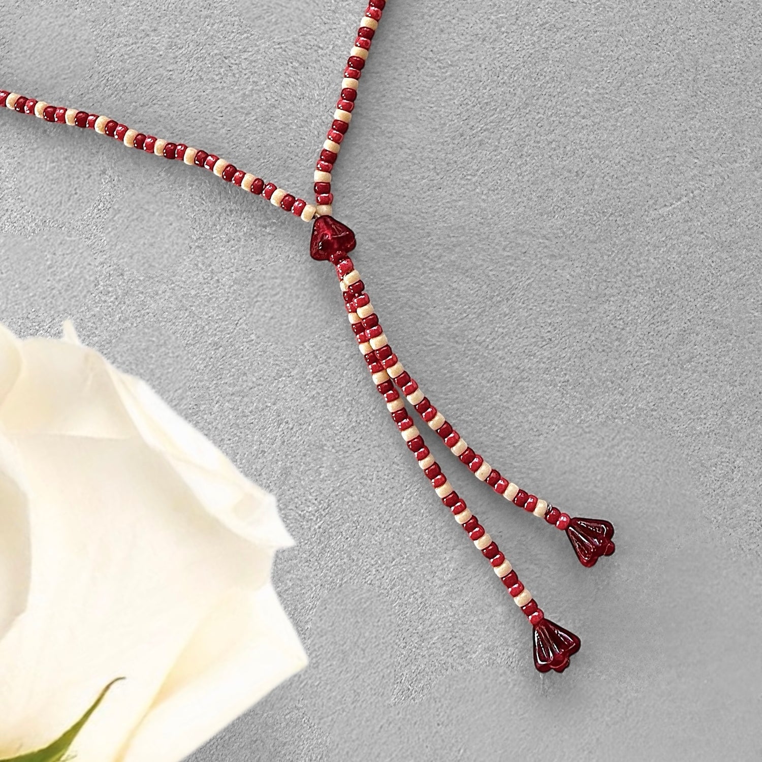Beaded necklace with red and white beads on a gray surface next to a white rose.