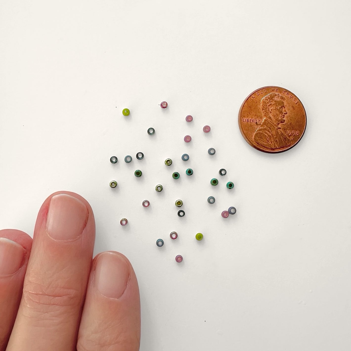 Size 11/0 Miyuki seed beads in multiple colors sprinkled on a white surface with human fingers and a penny in view for size reference