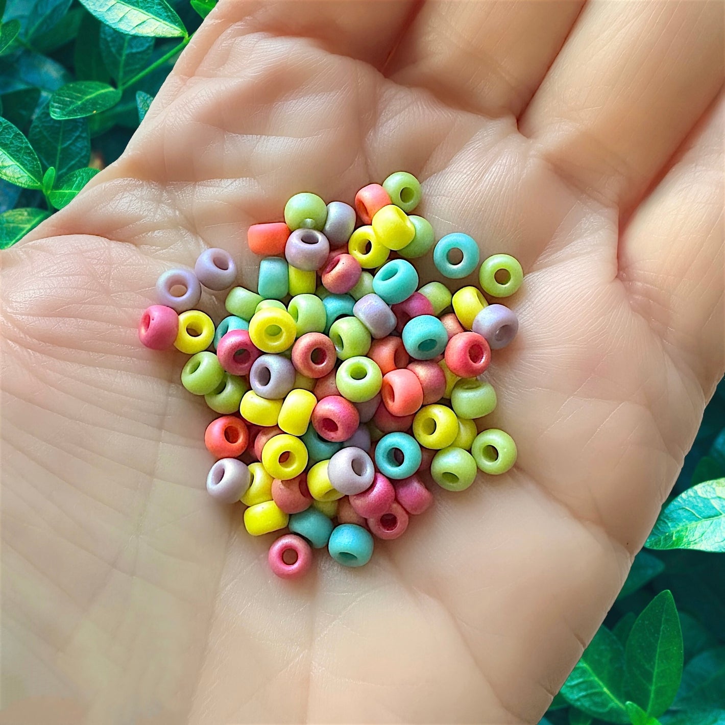 Human hand holding a colorful mix of Miyuki size 6/0 round rocaille seed beads with green leaves in the background