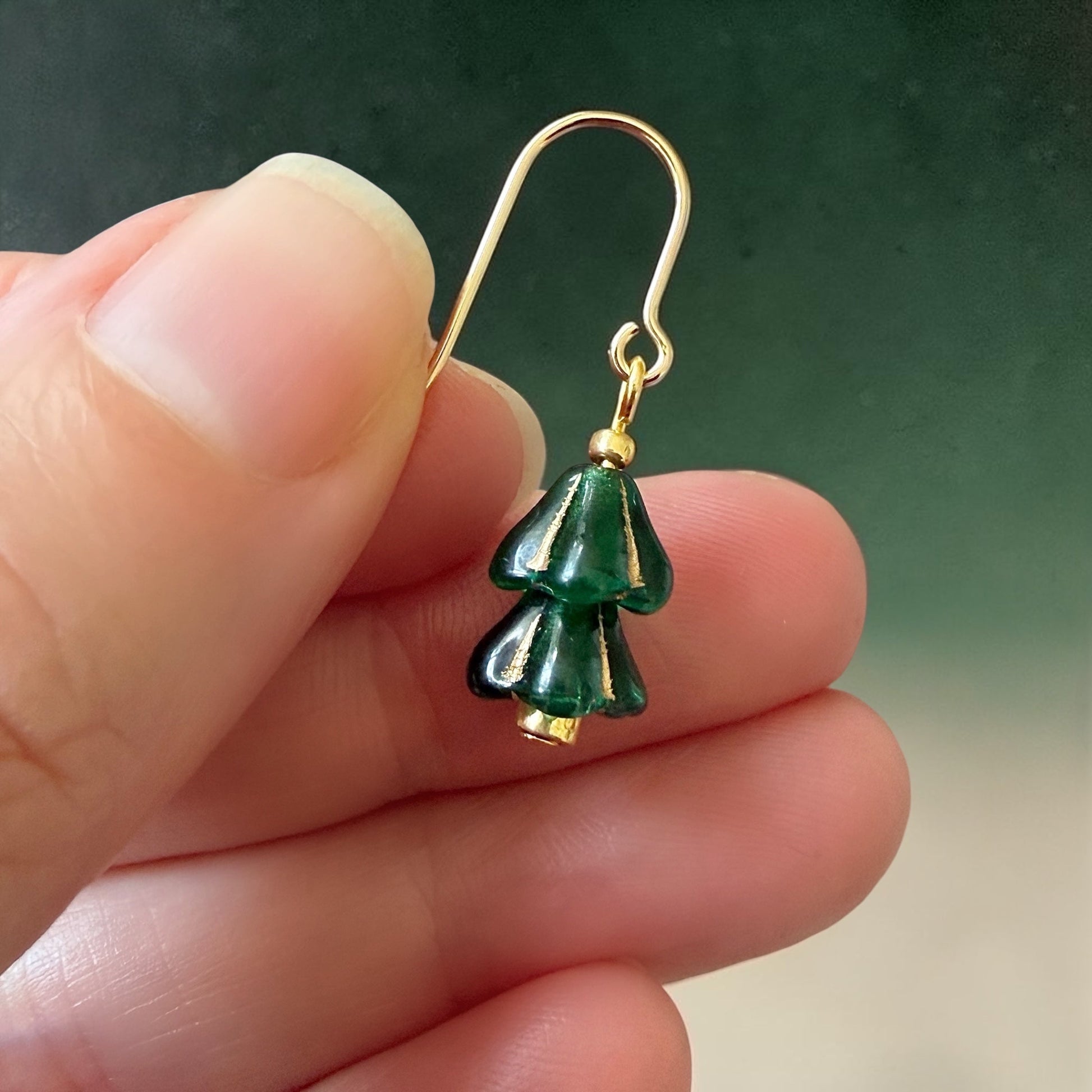 A woman's hand holding a green and gold beaded Christmas tree earring on a green gradient background.