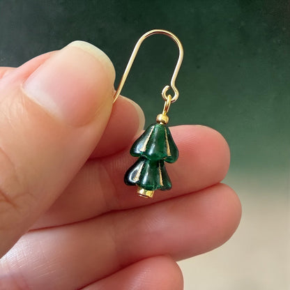 A woman's hand holding a green and gold beaded Christmas tree earring on a green gradient background.