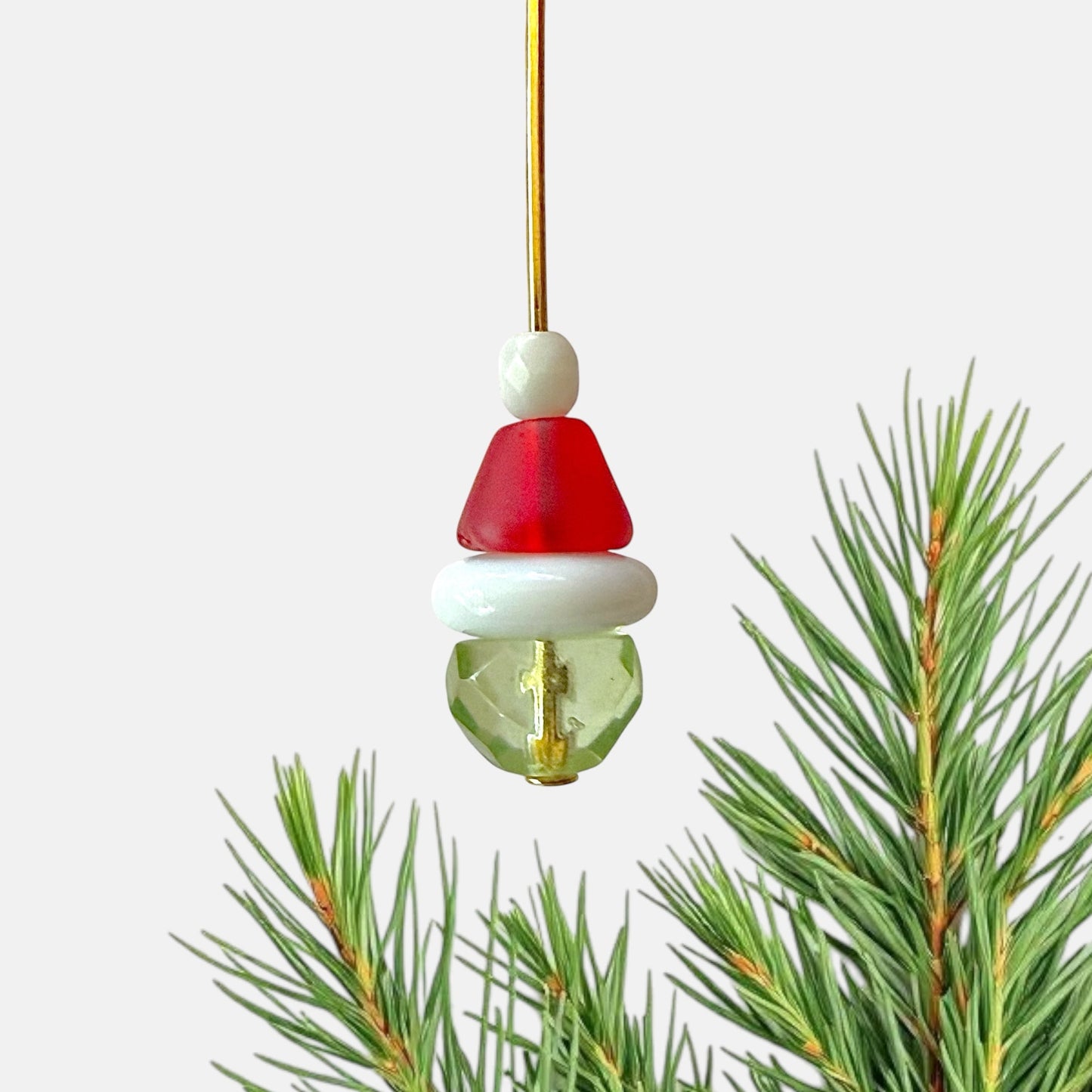 Beads assembled on a gold head pin to resemble the Grinch  with a white background and pine needles.