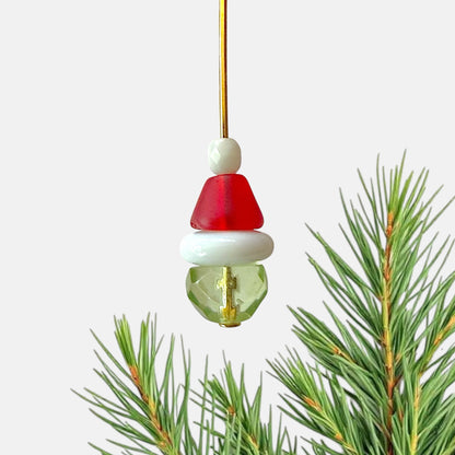 Beads assembled on a gold head pin to resemble the Grinch  with a white background and pine needles.