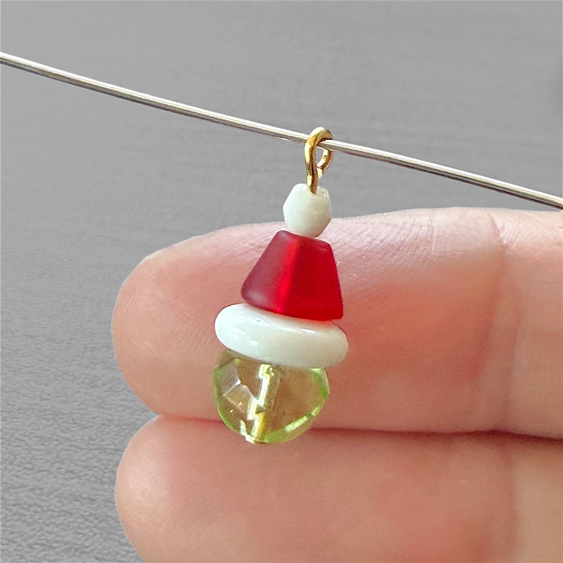 Fingers holding a beaded charm that resembles the grinch with a gold loop and a gray background.