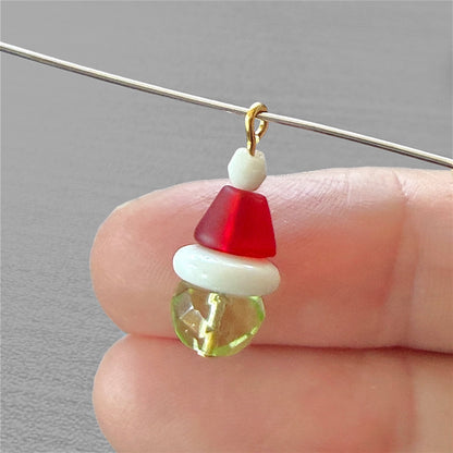 Fingers holding a beaded charm that resembles the grinch with a gold loop and a gray background.