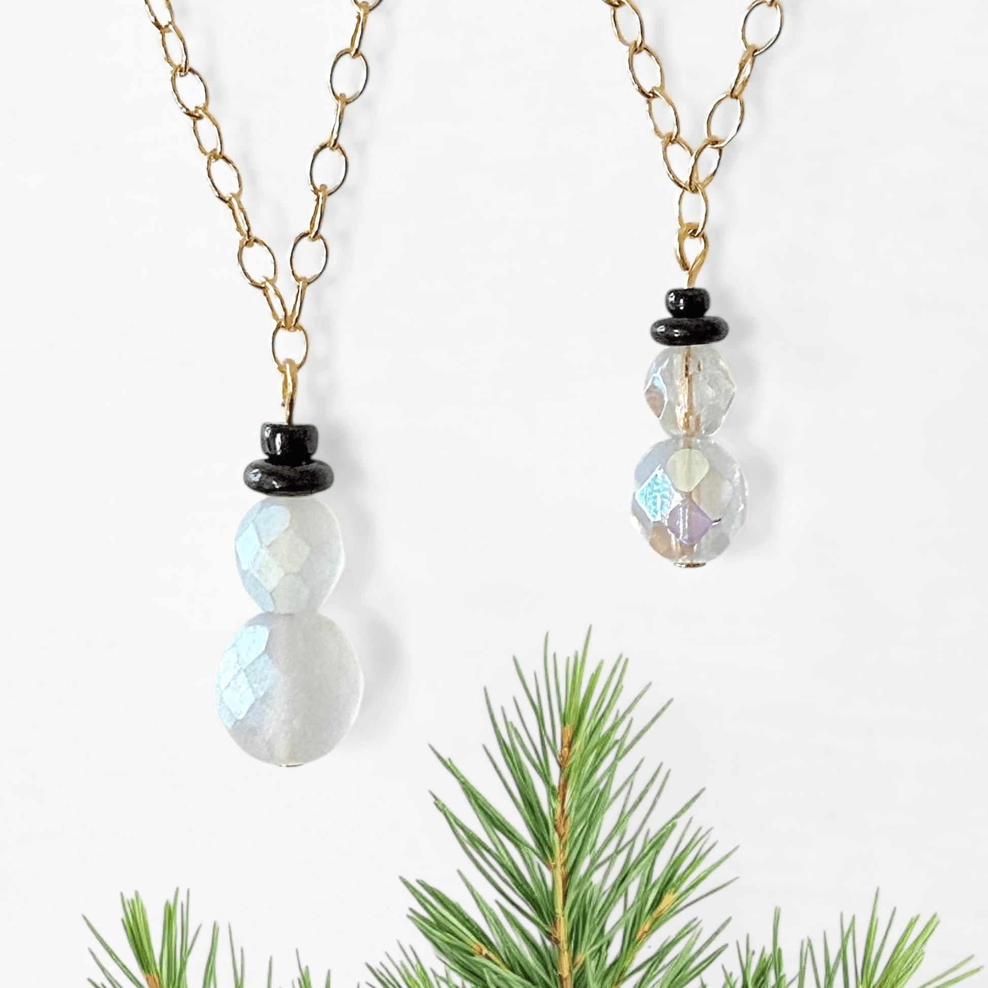 Two beaded snowman charms with white and crystal AB beads and black beads for the top hat attached to gold chains on a white background.