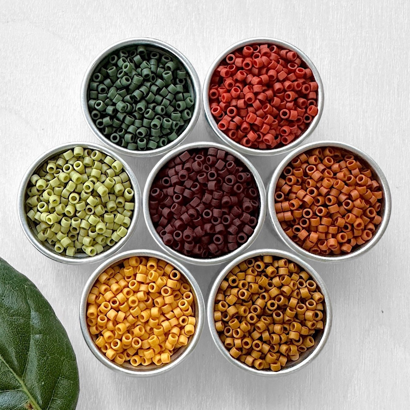 An arrangement of seven small containers of Miyuki 11/0 Delica beads in various autumn colors.