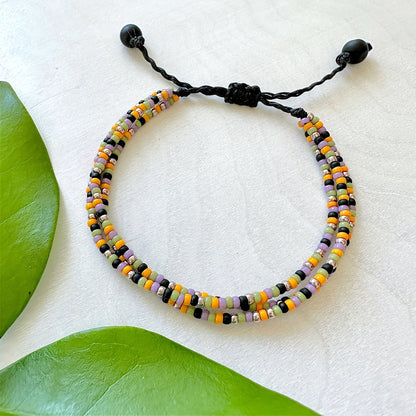 A three strand bracelet made with Miyuki round seed beads in faded Halloween colors with black cord on a white wood surface.