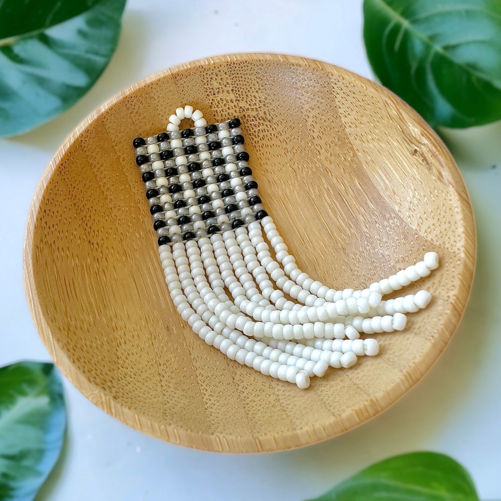 A beaded fringe earring with a black gingham pattern in a wooden dish on a white cloth