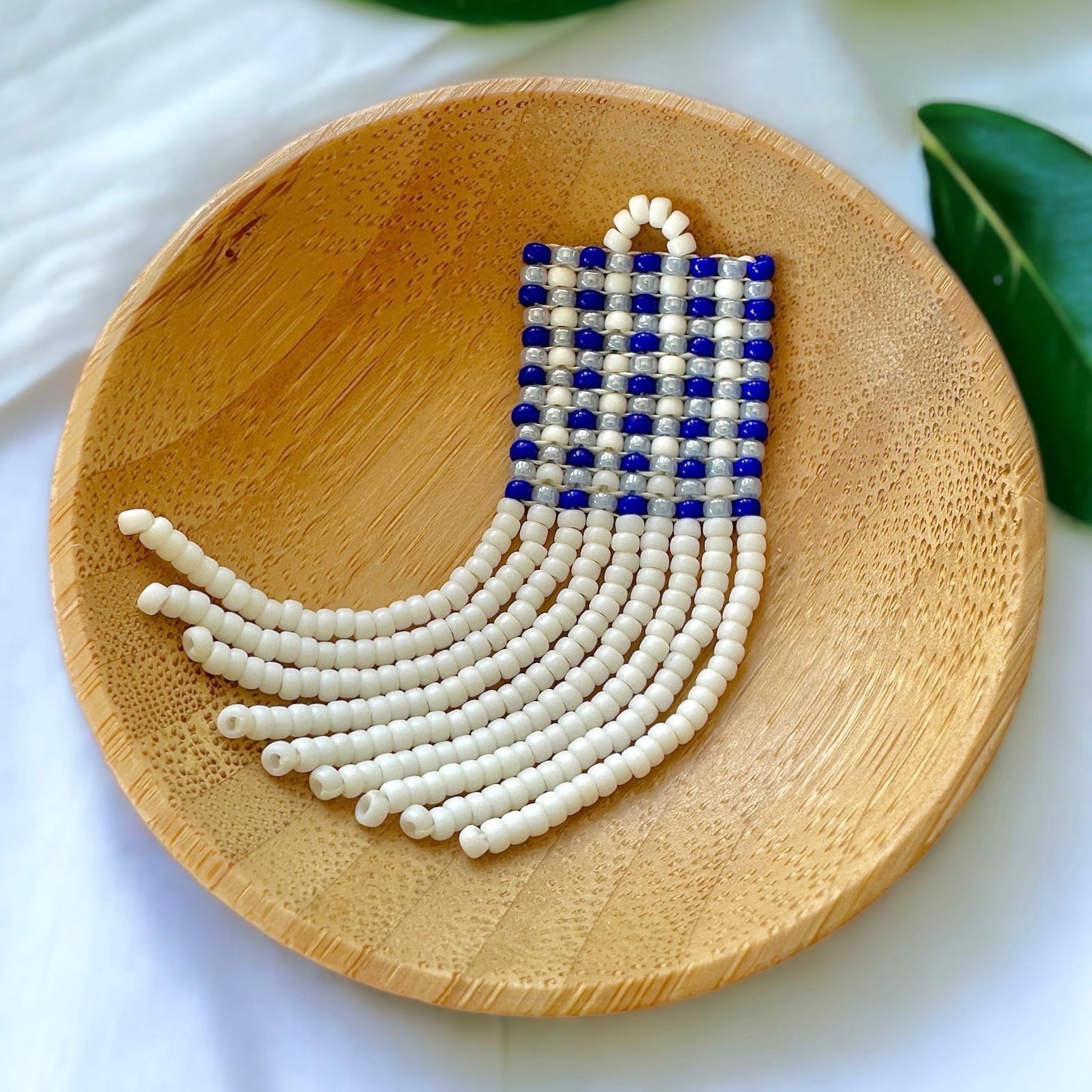 A beaded fringe earring with a blue gingham pattern in a wooden dish on a white cloth