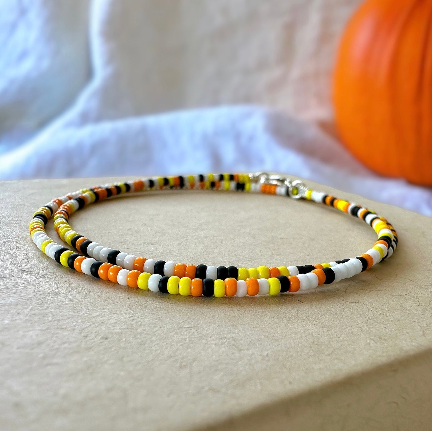 Candy corn dainty seed bead necklace made with black, white, yellow, and orange Miyuki 11/0 seed beads on a kraft box with a white cloth and orange pumpkin in the background