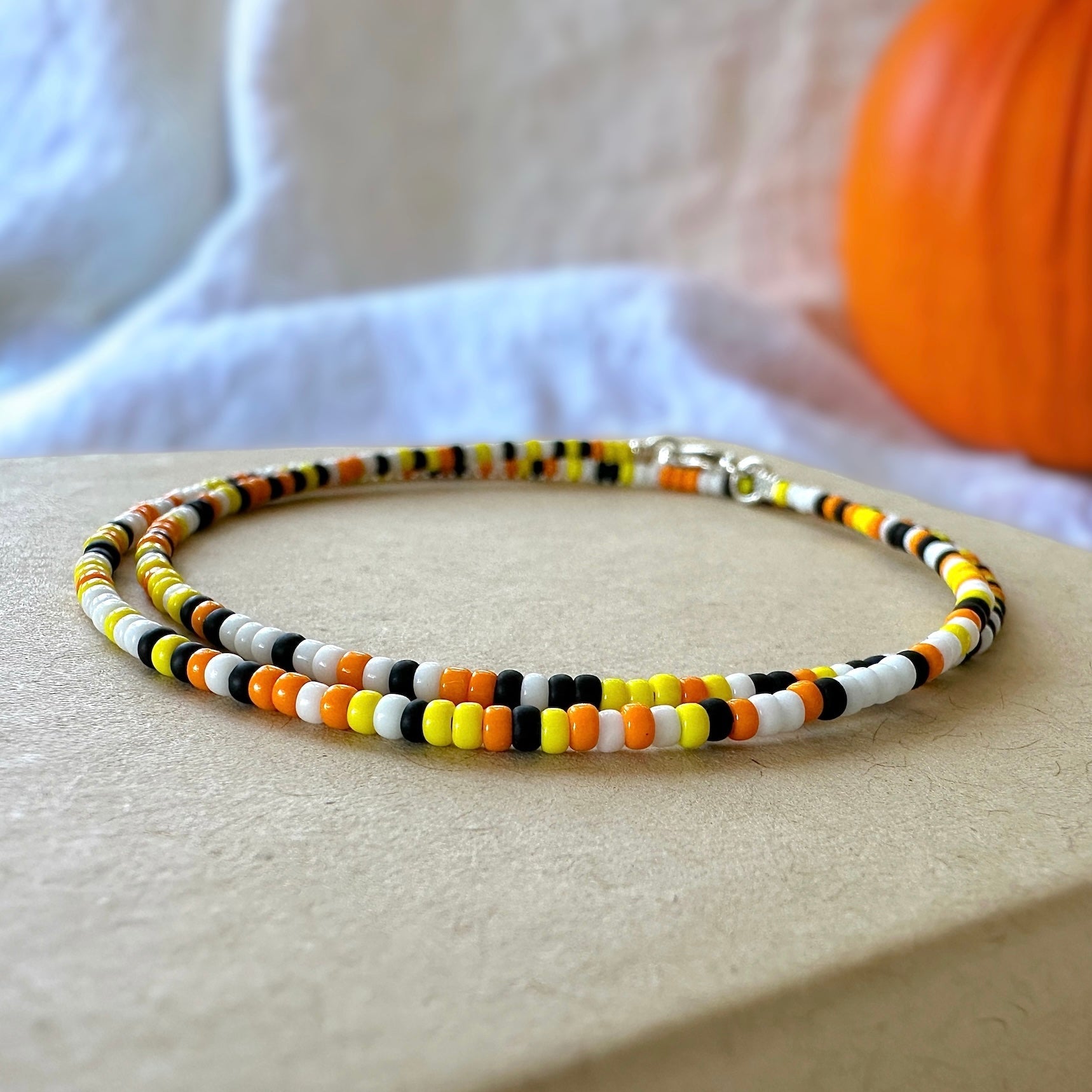 Candy corn dainty seed bead necklace made with black, white, yellow, and orange Miyuki 11/0 seed beads on a kraft box with a white cloth and orange pumpkin in the background
