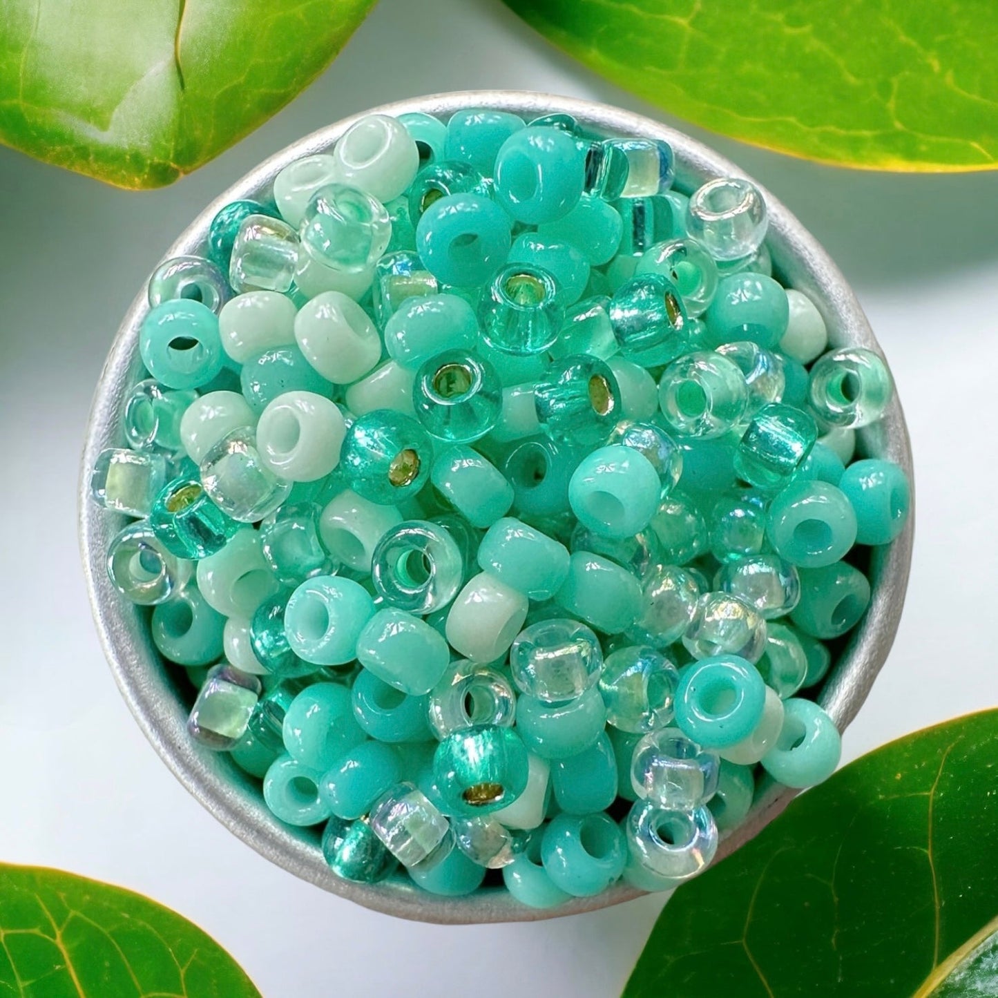 A mix of Miyuki round seed beads in shades of teal green, aqua, and mint green in a small metal container.