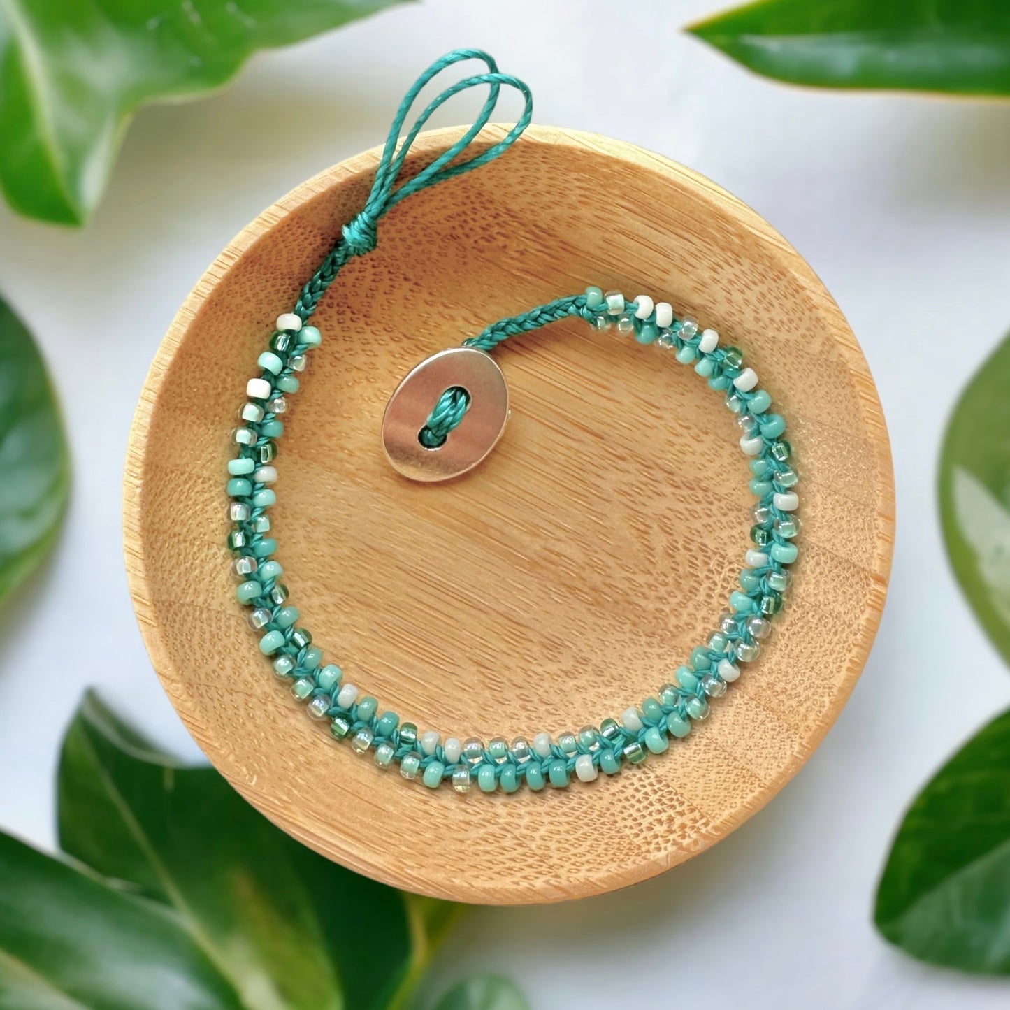 A beaded bracelet made with seed beads in shades of teal and mint green with a button closure in a wooden dish.