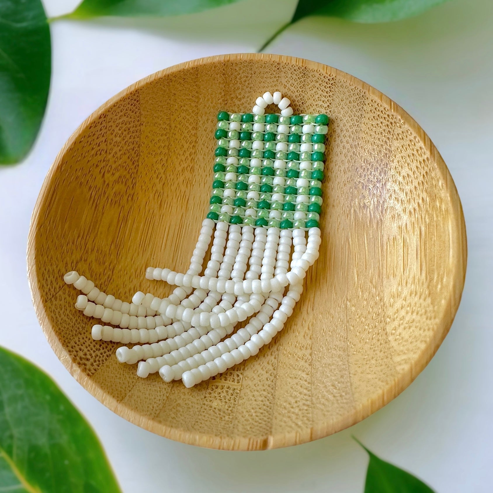 A beaded fringe earring with a green gingham pattern in a wooden dish on a white cloth