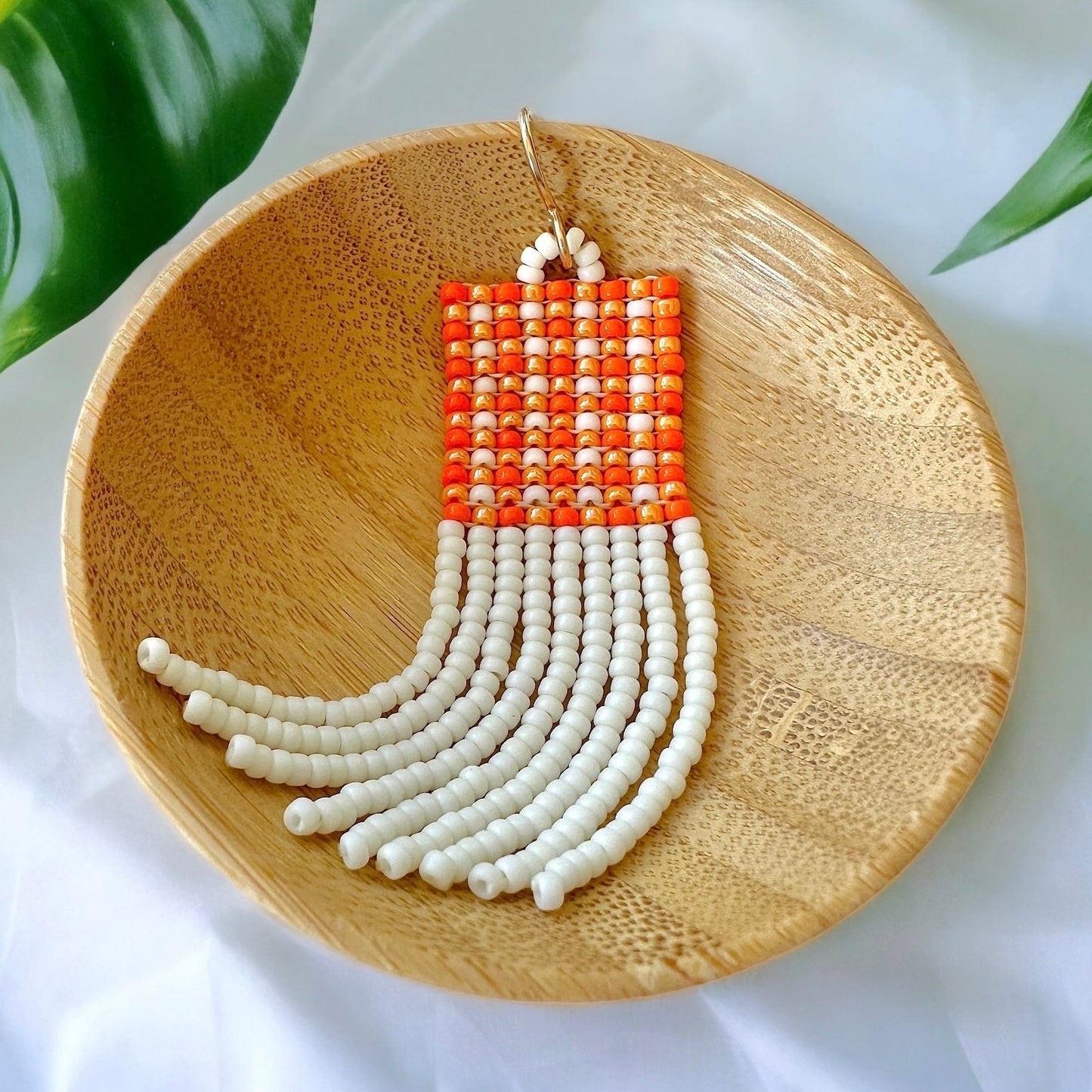 A beaded fringe earring with an orange gingham pattern in a wooden dish on a white cloth