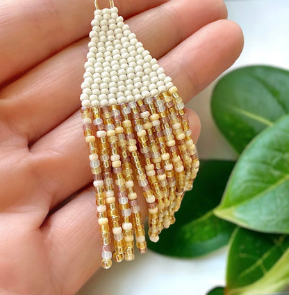 Brick stitch beaded fringe earring made with cream and golden topaz Miyuki 11/0 seed beads held in a woman's hand on a white background surrounded by green leaves