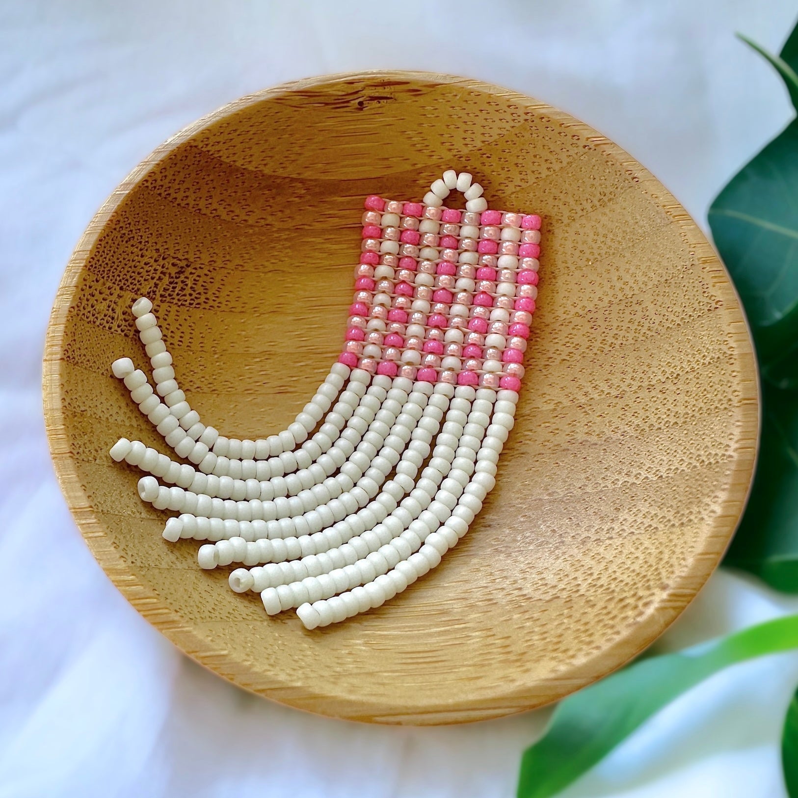A beaded fringe earring with a pink gingham pattern in a wooden dish on a white cloth